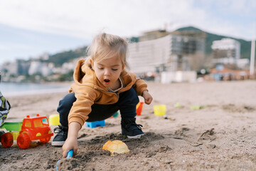 Little girl squats on the beach and digs the sand with her mouth open