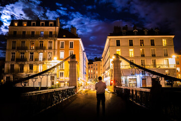 Grenoble, France - September 29, 2023: A man staying in the night in the middle of the Saint-Laurent footbridge - suspension bridge - now reserved for pedestrians.  - obrazy, fototapety, plakaty