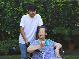 Obraz premium Portrait of Asian senior woman sitting on wheelchair, holding blue cup of tea or coffee in the garden, with taking care of her son.