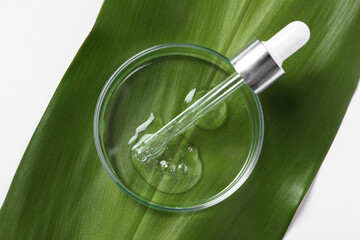 Petri dish with sample of cosmetic oil, pipette and green leaf on white background, top view