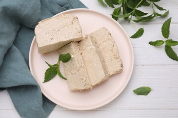 Plate with pieces of tasty halva and mint on white wooden table, flat lay