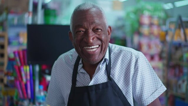 Joyful African American senior staff of grocery store standing by cashier checkout wearing apron and smiling at camera, portrait close-up with friendly charismatic expression