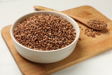 Bowl and spoon with dry buckwheat on white table, closeup