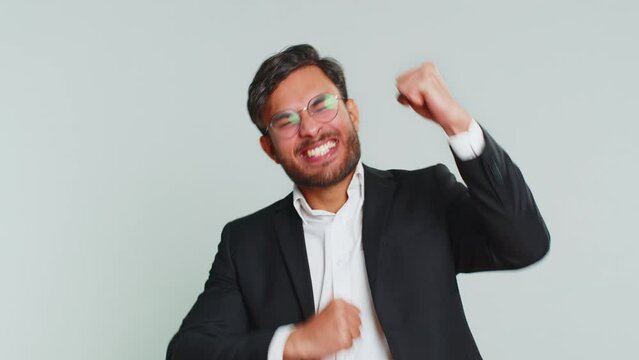 Overjoyed happy Indian businessman showing rock n roll gesture by hands, cool sign, shouting yeah with crazy expression, dancing, rejoicing victory win success. Male Arabian man guy on gray background