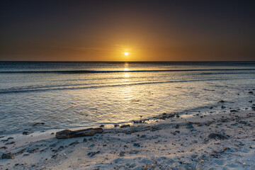 Sunset on the beach in Aruba. White sand littered with rocks, driftwood and shells. Gentle waves rolling towards shore. Setting sun in golden sky in the background. 
