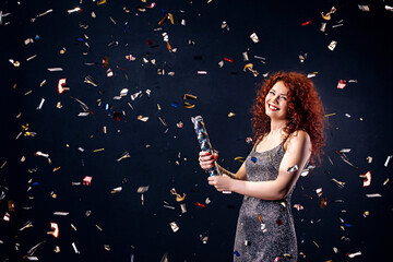 Red-haired, young woman in party  laughing while blowing up party popper isolated over black wall....