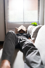Man with mental health problem lying on comfortable couch and covering face with book to sleeping - obrazy, fototapety, plakaty