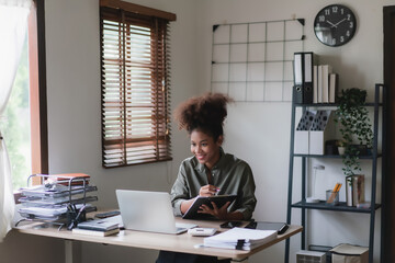 African american businesswoman reading finance business data on laptop and writing note in document