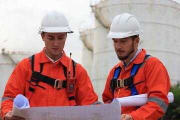 Team Engineers wearing safety harnesses working and discussing with blueprint at the Oil Refinery storage tanks. - obrazy, fototapety, plakaty