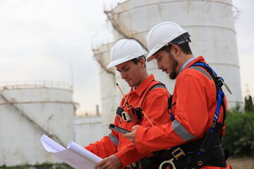 Team Engineers wearing safety harnesses working and discussing with blueprint at the Oil Refinery storage tanks.