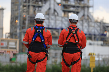 Back view of two engineers wearing fully safe uniforms, white hard hats, and safety harnesses with...