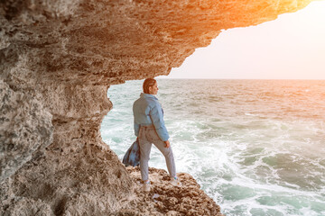 woman sea travel. A woman in a blue jacket stands on a rock above a cliff above the sea, looking at the stormy ocean. Girl traveler rests, thinks, dreams, enjoys nature