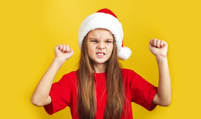 Angry little girl wearing Christmas hat standing isolated over yellow background wall in studio