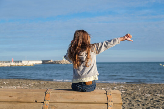 Ni&ntilde;a en la playa, vista de espaldas, octubre, pelo largo suelto, jugando en la arena, sentada mirando el mar, mano en alto signo mal, equivocado, 