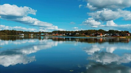 White cumulus clouds in a blue sky are beautifully mirrored in a blue lake against the background of an autumn forest and a suburban cottage village on a beautiful warm day. High quality FullHD