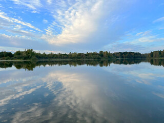 White cumulus clouds in a blue sky are beautifully mirrored in a blue lake against the background of an autumn forest and a suburban cottage village on a beautiful warm day. High quality FullHD