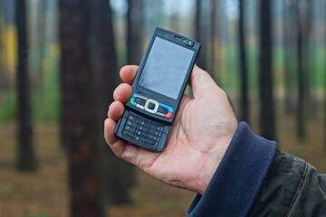 one old push-button mobile cell phone, black, plastic, small, compact, for calls and the Internet in the hand of a man on the street in the forest during the day