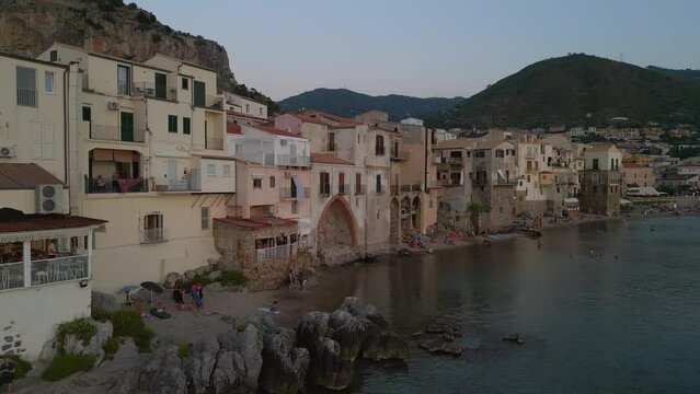 Drone tracks along seafront of Cefalu old port at Dusk. With restaurants overlooking the sea.  Beach, and mountains behind. People pack up to leave beach.