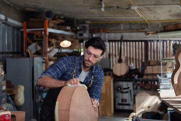 Woodworker in apron sanding the edge of guitar body using manual tool with sanding paper - obrazy, fototapety, plakaty