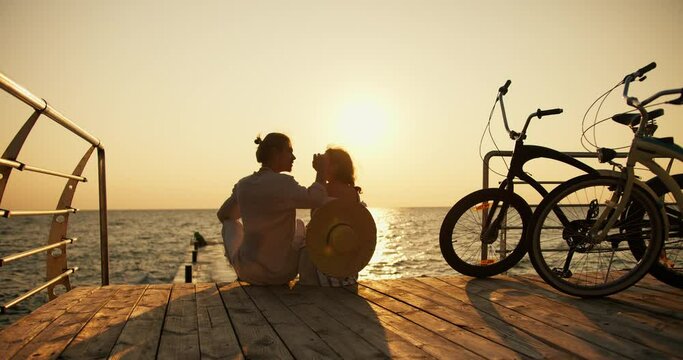 A happy couple, a guy in a light shirt and a girl in a straw hat, look at each other and sit on a beach covered with boards near the sea at sunrise. A guy and a girl came to the beach on bicycles