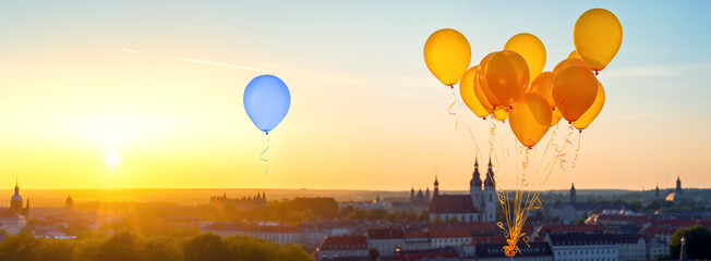 Standing out from the crowd concept. Blue balloon floating over a city separately from golden tied balloons during sunset - obrazy, fototapety, plakaty