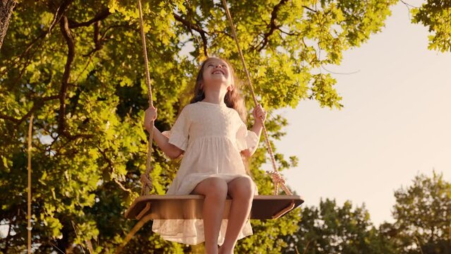 Child girl plays on wooden swing, dream fly. Happy little girl swings on swing in park under tree, sunset nature. Baby swing, kid girl smile in flight. Family happiness, dream, entertainment Concept