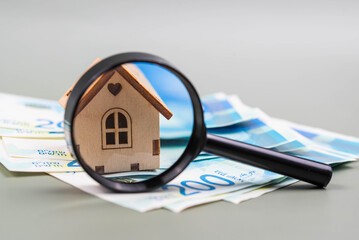 Buying a house, building repair and mortgage concept in Israel. Estimation real estate property with loan money and banking. Toy house with shekels cash banknotes under magnifying glass. - obrazy, fototapety, plakaty