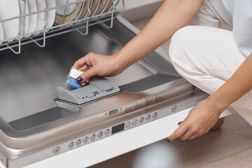 female hand puts dishwasher tablet into open automatic built-in dishwasher machine with dirty dish...