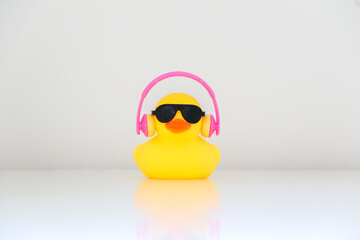 A little cool and hip yellow rubber duck wearing black sunglasses and pink headphones in the middle of white table background, listening to music. Be cool, be smart, be hip, music concept.