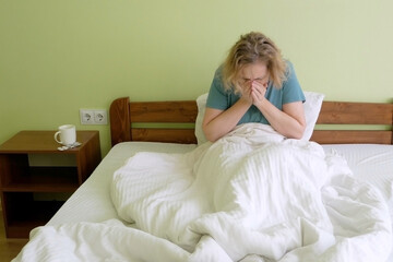 Middle aged woman sit in bed under warm blanket blow running nose using tissue, and coughs,   sick female suffer from influenza or grippe at home