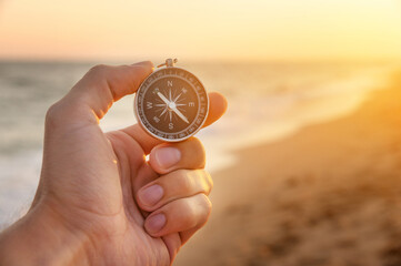 man's hand holds a compass on the beach. Hand with compass over sand by the sea, direction and adventure concept - obrazy, fototapety, plakaty