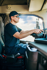 Handsome experienced male truck driver with a hat sitting and driving his truck. Professional transportation and truck drivers concept. Side view.