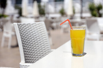 Glass of orange juice with plastic straw on white table in restaurant outdoor lounge zone in sunny weather