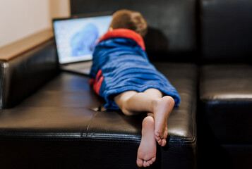 A boy child in pajamas lies, rests on a black leather sofa, watching a movie on a laptop online via...