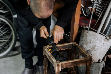 An adult male professional woodworker, carpenter, worker glues a chair, repairs, restores furniture indoors, workshop, clamping wooden products with a clamp. Close-up photography, work concept.