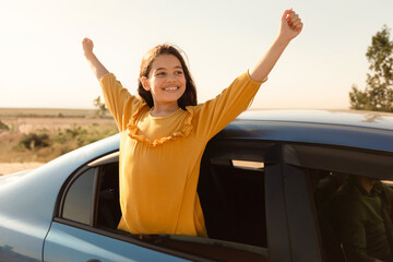 Joyful girl with arms raised from car, sunny day