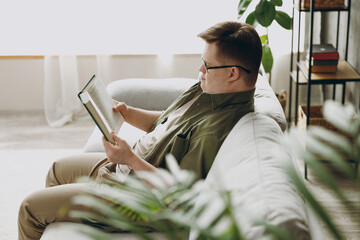 Side view young calm man with down syndrome wears glasses casual clothes reading book sits on grey sofa couch stay at home flat rest spend free time in living room. Genetic disease world day concept.