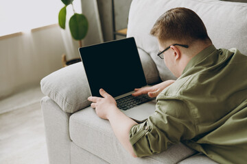 Young IT man with down syndrome wears casual clothes work use blank screen laptop pc computer lay on grey sofa couch stay at home rest spend free time in living room Genetic disease world day concept