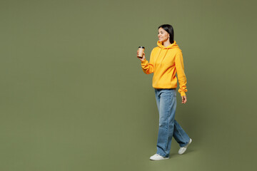 Full body young Latin woman wearing yellow hoody casual clothes hold takeaway delivery craft paper brown cup coffee to go isolated on plain pastel green background studio portrait. Lifestyle concept.