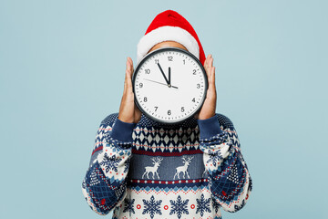 Young man wearing knitted sweater Santa hat posing holding in hand cover face with clock isolated on plain pastel blue cyan color background. Happy New Year 2024 celebration Christmas holiday concept. - obrazy, fototapety, plakaty