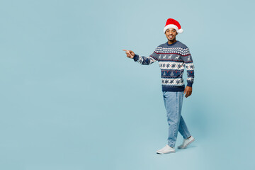 Fototapeta na wymiar Full body side view young man wear knitted sweater Santa hat posing walk go strolling point aside on area isolated on plain blue background. Happy New Year 2024 celebration Christmas holiday concept.