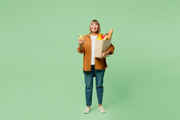 Full body elderly woman wear brown shirt casual clothes hold shopping paper bag with food product use mobile cell phone surfing internet isolated on plain green background. Delivery service from shop.