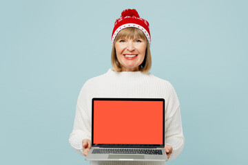 Merry elderly IT woman 50s years old wear sweater red hat posing hold use work on blank screen area laptop pc computer isolated on plain blue background. Happy New Year celebration Christmas concept.