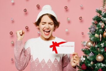 Merry young woman in white sweater hat posing hold store gift certificate coupon voucher card do...