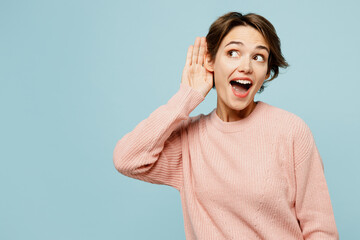 Young curious nosy woman wear beige knitted sweater casual clothes try to hear you overhear listening intently isolated on plain pastel light blue cyan background studio portrait. Lifestyle concept.