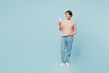 Full body young smiling happy woman wear beige knitted sweater casual clothes hold in hand use mobile cell phone chatting online isolated on plain pastel light blue cyan background. Lifestyle concept.