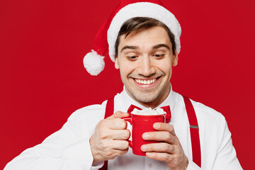 Merry young man wears white shirt Santa hat posing hold in hand cup with cocoa and marshmallow isolated on plain red background studio portrait. Happy New Year Christmas celebration holiday concept. - Powered by Adobe
