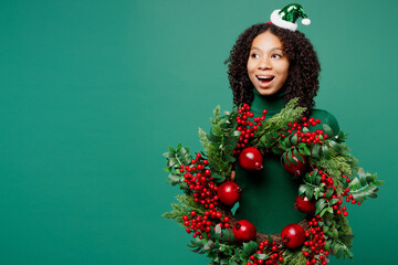 Merry fun little kid teen girl wear hat casual clothes posing hold Christmas handmade wreath look aside isolated on plain green background studio portrait. Happy New Year celebration holiday concept.