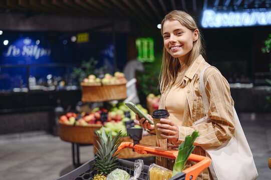 Young happy customer woman wear casual clothes use mobile cell phone drink coffee tea shopping with cart at supermaket store grocery shop buy choose products in hypermarket. Purchasing food concept.