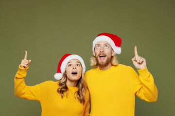 Merry shocked fun young couple two friends man woman wearing sweater Santa hat posing point index finger overhead on area isolated on plain green background. Happy New Year Christmas holiday concept.